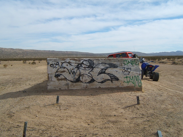 Graffiti on concrete barrier in desert landscape. Blue quad bike and red SUV visible in background