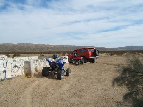 1988 Ford Bronco - A red 1988 Ford Bronco and two ATVs are parked in a dry, desert landscape next to a graffiti-covered concrete barrier.