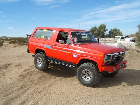 1988 Ford Bronco - A red Ford Bronco is parked on a sandy surface with a clear blue sky above.
