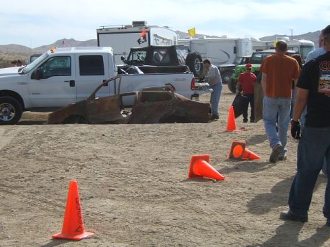 1988 Ford Bronco - A heavily damaged car chassis sits in a dirt area, with several trucks and trailers in the background.