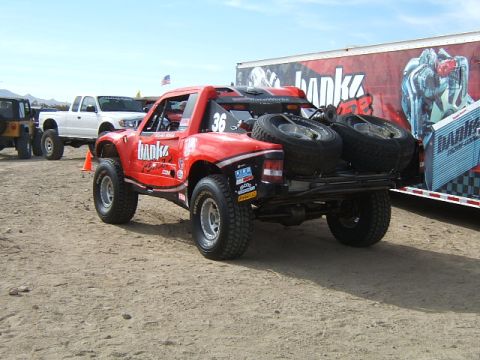 1988 Ford Bronco - Off-road race truck with spare tires and sponsor decals parked next to a trailer. Other vehicles are in the background.
