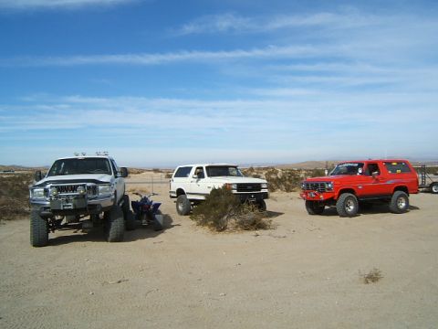 1988 Ford Bronco - Three modified SUVs and an ATV are parked in a dry, dusty landscape under a blue sky with scattered clouds.