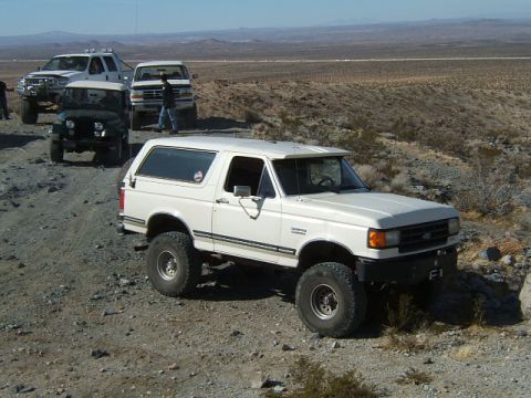 1988 Ford Bronco - A group of off-road vehicles are parked on a dusty trail in a desert landscape.