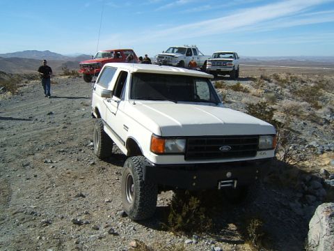 1988 Ford Bronco - Several trucks and a white Ford Bronco are parked on a dusty, off-road trail with mountains in the background.
