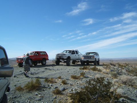 1988 Ford Bronco - Several off-road vehicles are parked on a dusty, rocky terrain with a vast, arid landscape in the background.