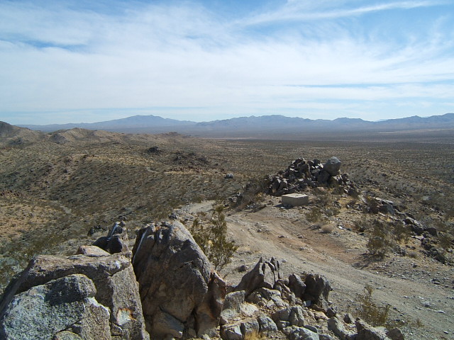 Desert landscape with rocky foreground, sparse vegetation, a small building on a hill, and distant mountains under a cloudy sky