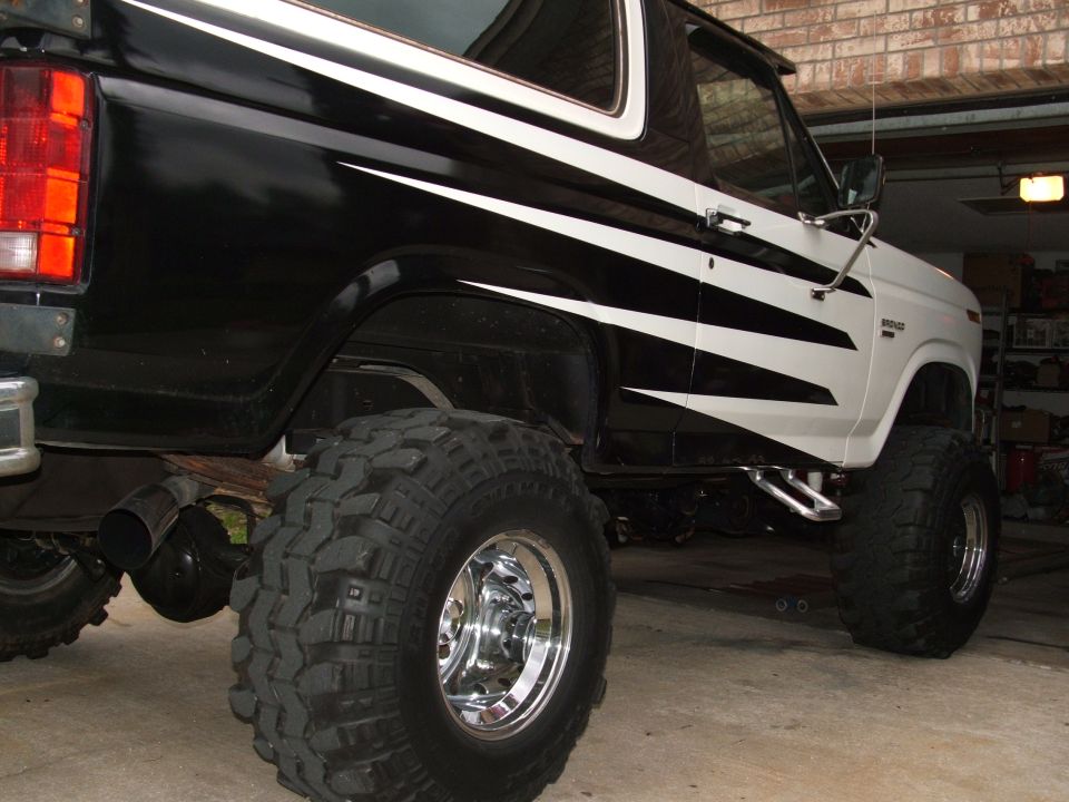 Lifted 1985 Ford Bronco with black and white graphics, large off-road tires, and chrome wheels