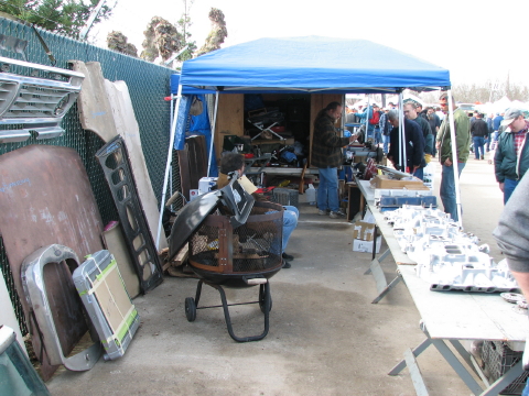 1973 Ford Ranchero - A swap meet vendor stall under a blue pop-up canopy with used auto parts spread across folding tables and stacked on the pavement, includin…