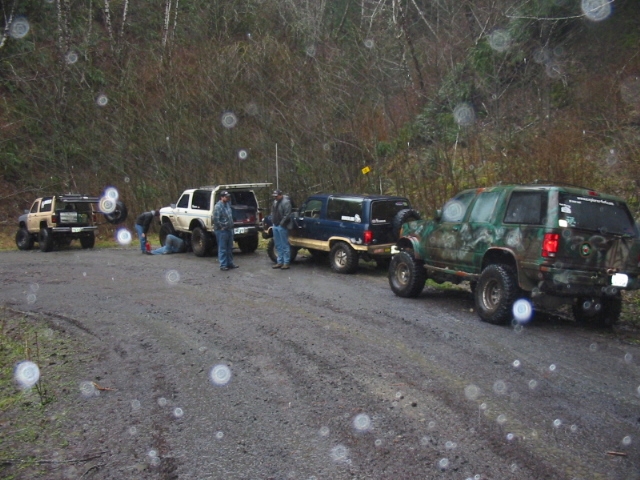 Several modified SUVs and trucks parked on a muddy dirt road, with people around them