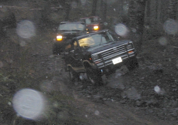 Three black SUVs driving on a wet, muddy trail in the woods