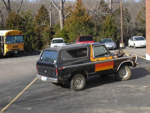 1979 Ford Bronco - A black and orange Ford Bronco is parked in a lot with its hood up, alongside other vehicles and a school bus.