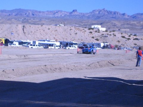 1990 Ford Bronco - blue off-road truck rounds a dirt track in front of a paddock of RVs and trucks as a red-vested flagger watches trackside.
