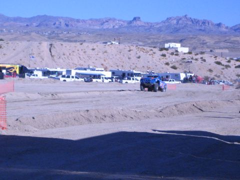 1990 Ford Bronco - blue off-road truck drives a dusty dirt course in front of parked RVs and pickups under rugged desert mountains.