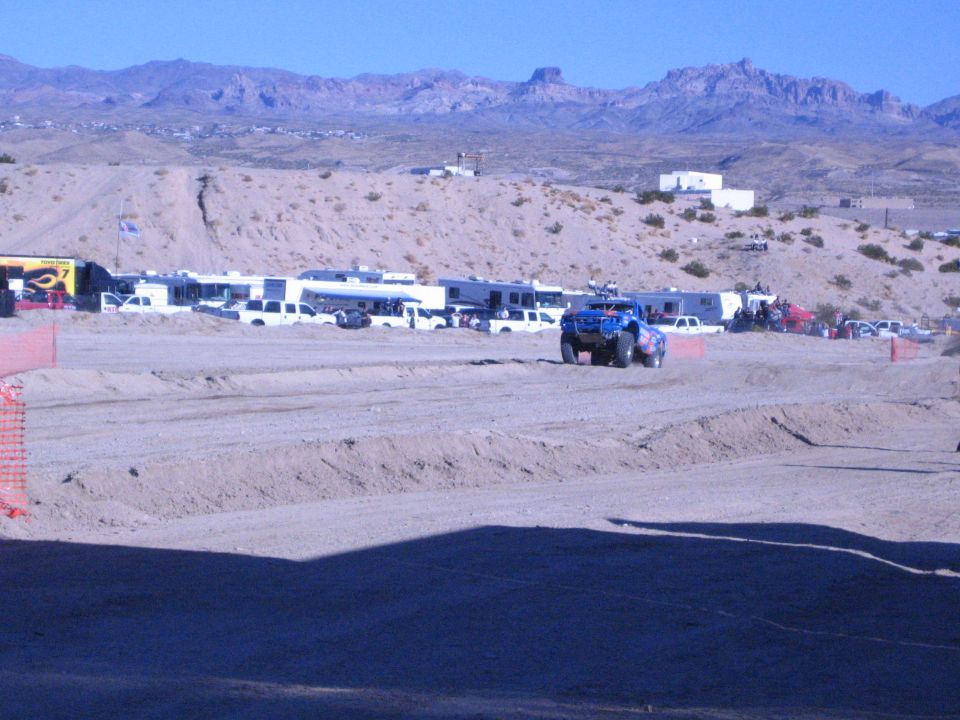 Blue off-road truck on a desert dirt course past parked RVs and pickups