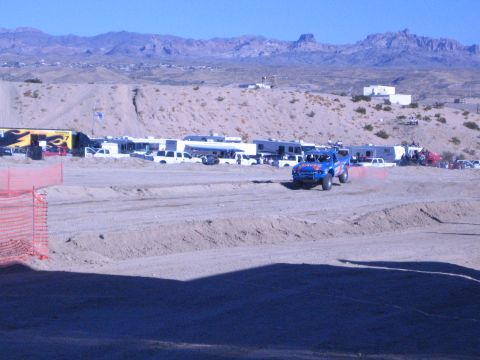 1990 Ford Bronco - blue off-road race truck runs a graded dirt course bordered by orange safety fencing with RVs and mountains behind.