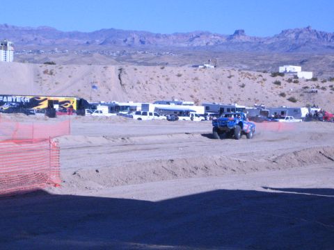 1990 Ford Bronco - blue off-road truck drives a dirt desert course past a yellow Fabtech banner with orange fencing and RVs in the paddock.
