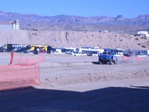 1990 Ford Bronco - blue off-road truck kicks up dust on a dirt desert course beside orange fencing and a Fabtech banner with a casino building behind.