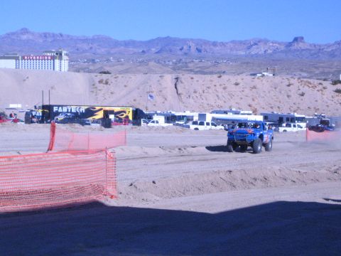 1990 Ford Bronco - blue off-road race truck kicks up dust on a desert dirt course, with a FABTECH trailer, RVs and a hillside casino in the background.
