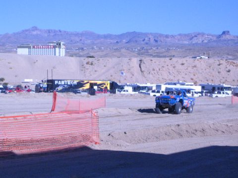 1990 Ford Bronco - lone blue off-road race truck drives a desert dirt track past an orange safety fence, with pit RVs and a casino on the hillside behind.