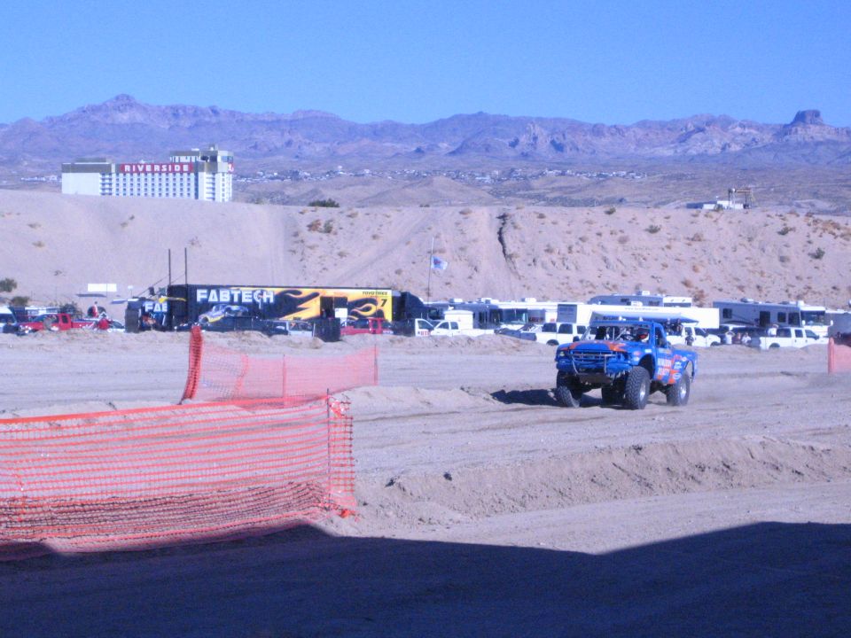 Blue off-road race truck on desert dirt track near orange safety fence and pit area