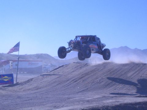 1990 Ford Bronco - open-cockpit off-road buggy catches air off a dirt jump in the desert, with a US flag and Sunoco banner along the course.