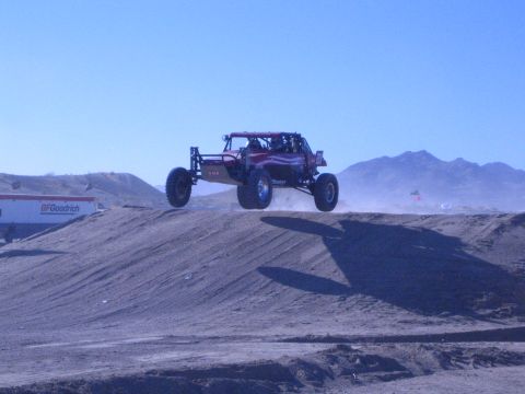 1990 Ford Bronco - dark red tube-chassis off-road buggy flies over a dirt jump in the desert, with a BFGoodrich banner along the track.