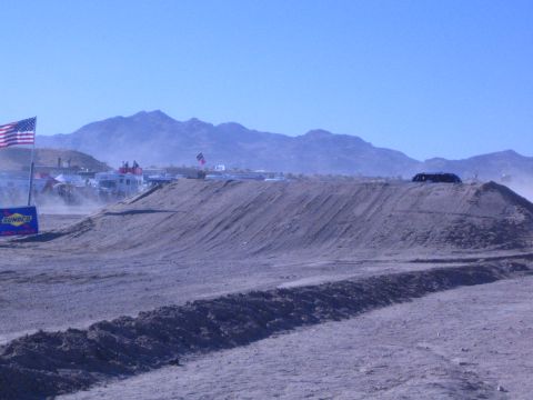 1990 Ford Bronco - desert dirt race course with a sculpted berm, US flag and Sunoco banner, with pit RVs and vehicles lined up in the distance.