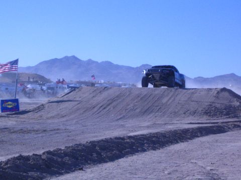 1990 Ford Bronco - dark off-road race truck crests a dirt berm in the desert, kicking up dust beside a US flag and Sunoco banner.