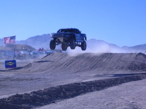 1990 Ford Bronco - blue off-road race truck launches off a dirt jump in the desert, with a US flag and Sunoco banner lining the course.