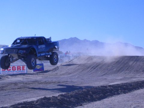 1990 Ford Bronco - blue off-road race truck marked Riviera flies off a jump past a SCORE International banner at a dusty desert event.