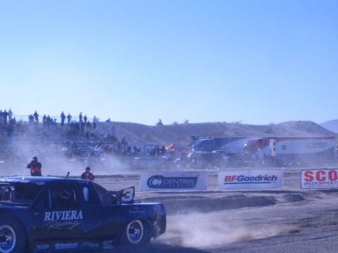 1990 Ford Bronco - blue Riviera Racing number 3 off-road truck runs a dusty dirt course in front of grandstands and BFGoodrich and SCORE banners.