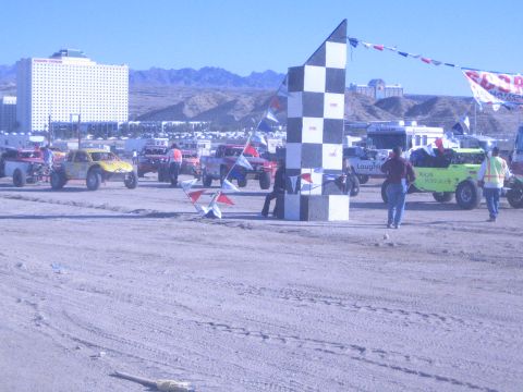 1990 Ford Bronco - checkered start tower stands in a dusty desert staging area surrounded by off-road race trucks, flags, crew and a hillside casino.