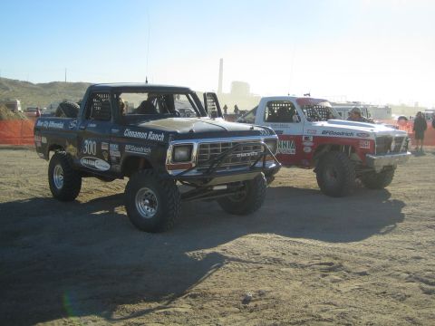 1990 Ford Bronco - black vintage Ford-style off-road truck lettered Cinnamon Ranch number 300 sits staged beside another race truck in the desert at golden ho…
