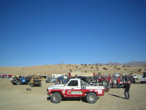 1990 Ford Bronco - white-and-red number 301 off-road race truck sits among rows of staged race vehicles in a sandy desert lot under clear blue sky.