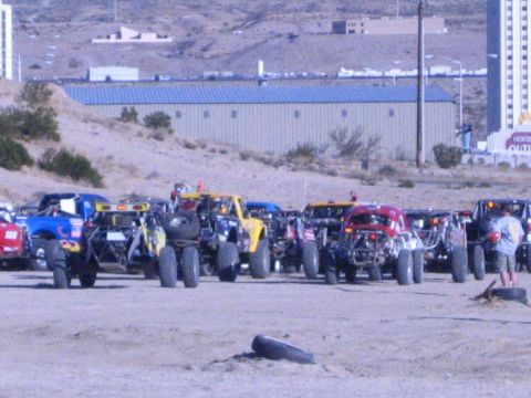 1990 Ford Bronco - row of off-road race trucks and buggies sits packed together in a dirt staging lot in front of a long blue metal building and hills.
