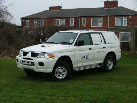 2006 Mitsubishi L200 - A white SUV with BT branding is parked on a grassy lawn in front of a brick house.