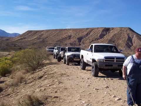1981 Ford F-150 - A convoy of off-road vehicles travels down a dusty desert trail under a clear blue sky.