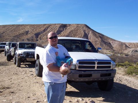1981 Ford F-150 - A man holds a baby in front of a white pickup truck, with other off-road vehicles lined up behind.