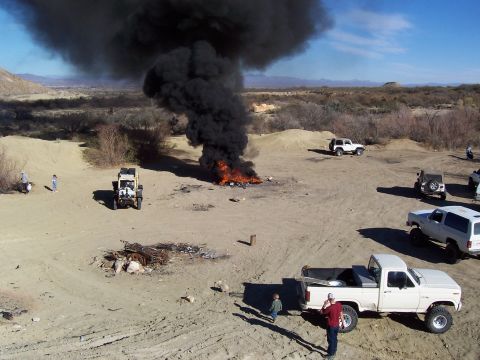 1981 Ford F-150 - A large fire billows black smoke into the sky in a desert area, with several vehicles and people observing the scene.