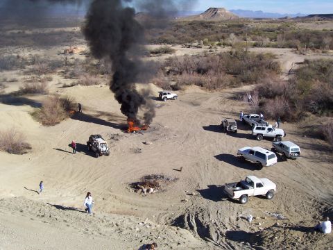 1981 Ford F-150 - Several off-road vehicles are gathered in a desert area with a large fire producing black smoke.