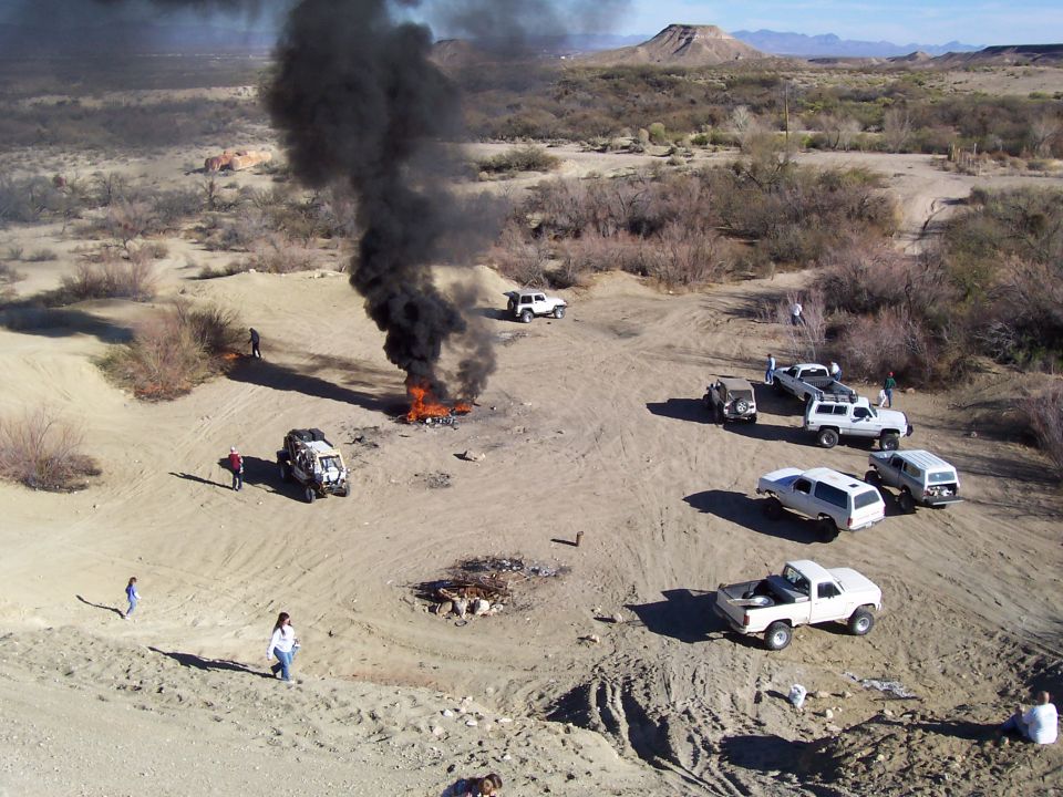 1981 Ford F-150 pickup truck, white, with a fire and black smoke nearby, surrounded by other off-road vehicles in a desert landscape
