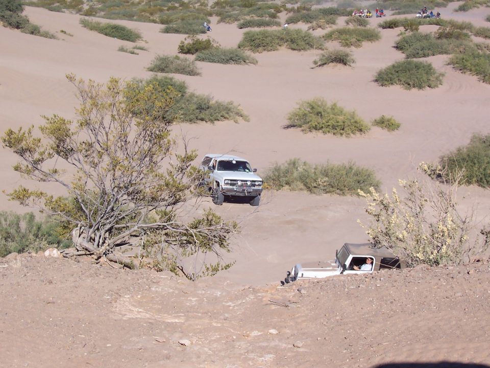 White pickup truck with camper shell driving on sand dunes, another truck partially buried