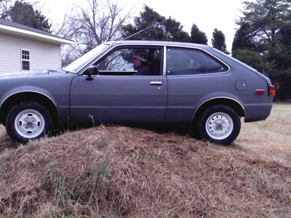 Gray 1979 Honda Accord coupe with white wheels, driver wearing a hat, parked on a dirt mound next to a house