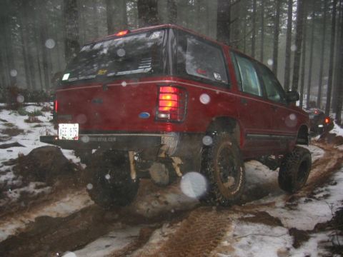 1991 Ford Explorer - A red 1991 Ford Explorer navigates a challenging off-road trail in a wooded area with snow and mud.