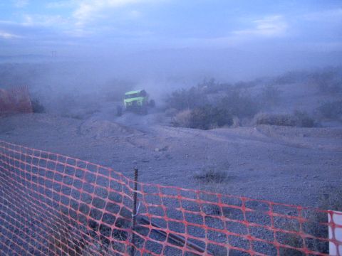 Green off-road buggy climbing dusty desert track behind orange safety fence
