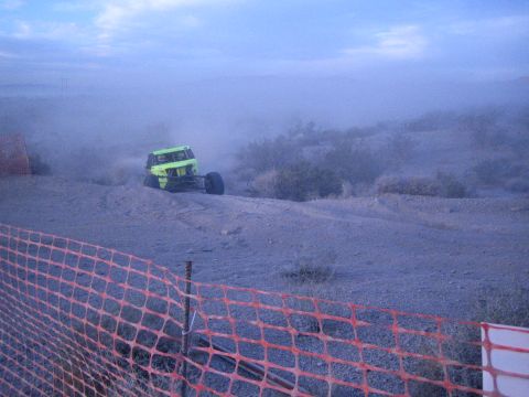 Green tube-chassis off-road buggy on desert berm behind orange safety fence