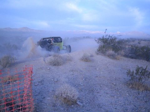 Green off-road buggy throwing dust cresting desert rise past orange fence