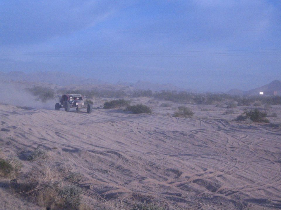 Red open-wheel off-road buggy on sandy desert flat with mountains behind