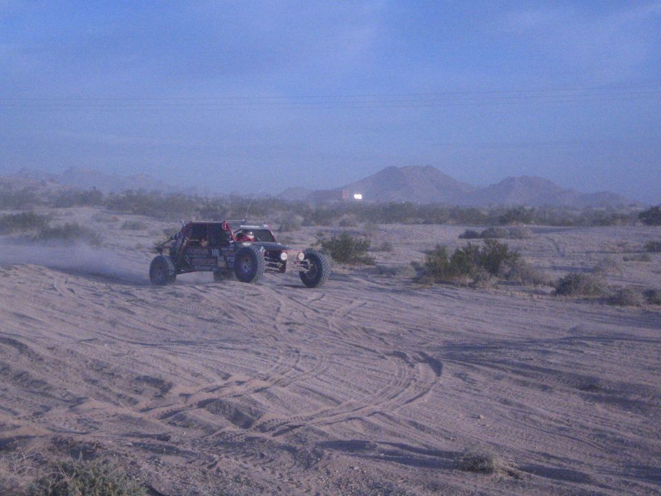 Open-wheel off-road buggy turning on sandy desert flat with mountains behind
