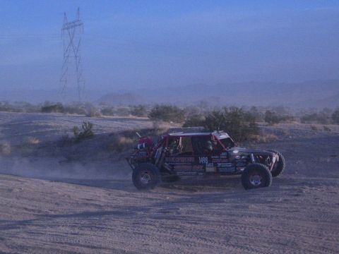 Off-road race buggy number 1498 on dusty desert track with power tower behind